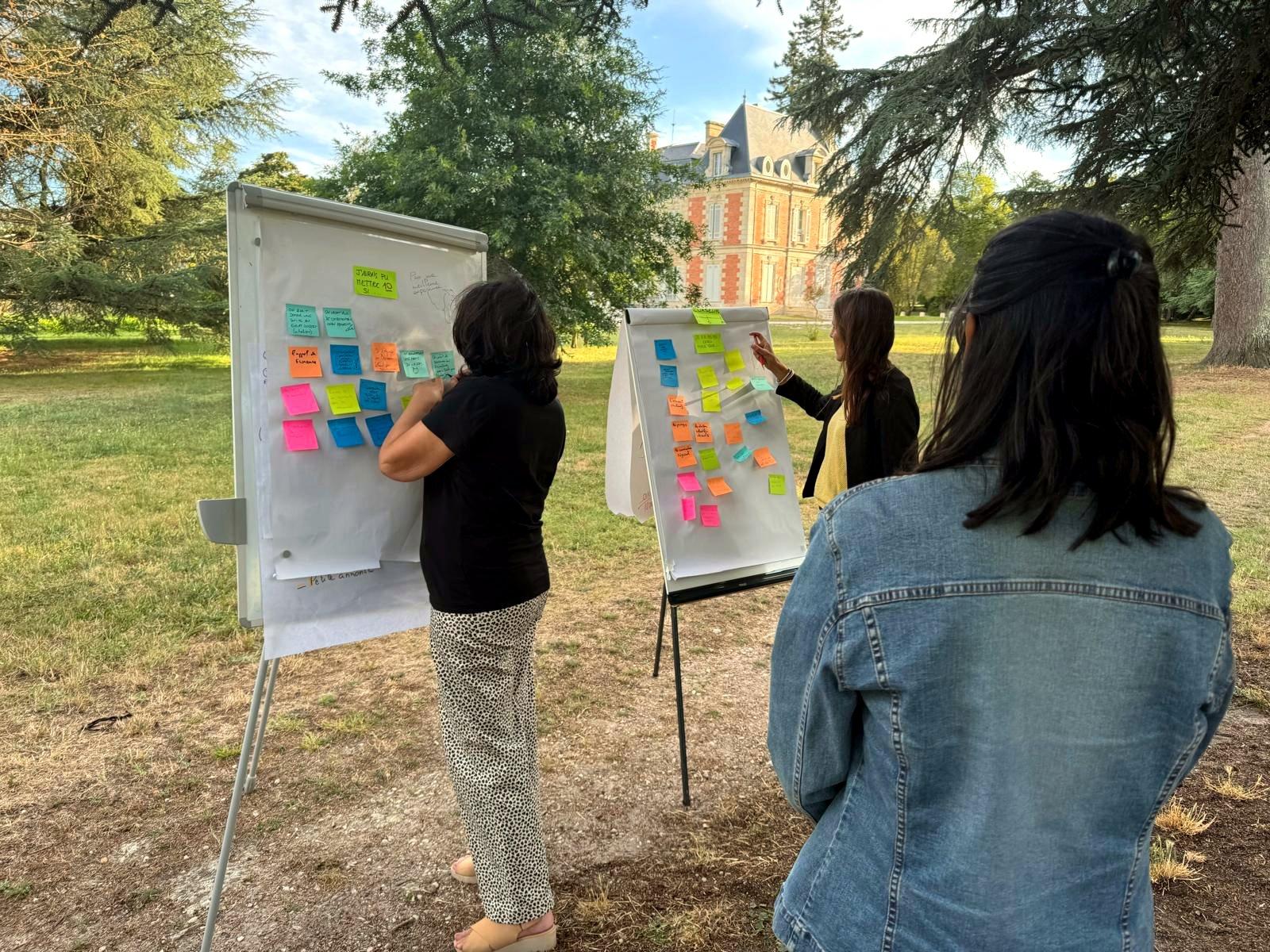 Trois personnes de dos travaillent sur des paperboards couverts de post-it, dans le parc d'un château.