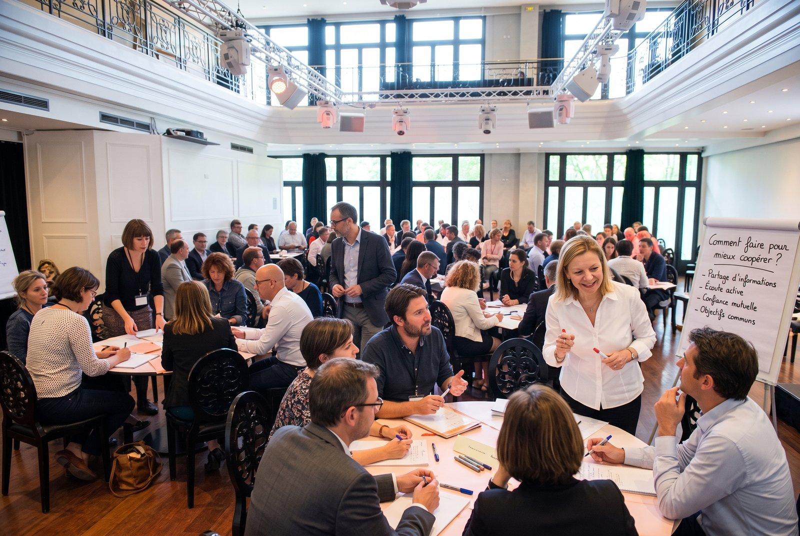 Une grande salle de séminaire où de nombreux participants travaillent en petits groupes autour de tables rondes. À droite, un paperboard titré « Comment faire pour mieux coopérer ? ».